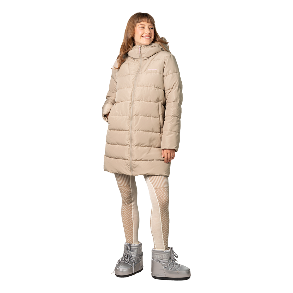 En kvinde iført en lang, beige dynejakke med hætte står afslappet og smilende. Jakken er tætsyet i vandrette baner og når til knæene. Hun har mønstrede, lyse strømpebukser og sølvglinsende Moon Boot-støvler, der giver et vinterligt udtryk. Hendes hænder er i lommerne, og helhedsindtrykket er varmt, moderne og funktionelt til kolde dage.