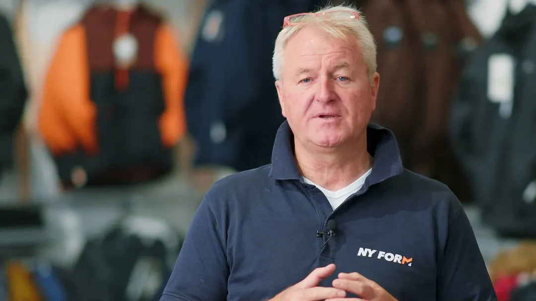 En ældre mand står midt i billedet og taler direkte til kameraet. Han har lyst hår, briller hvilende på hovedet og bærer en mørk polo med logoet "NY FORM". En lille clip-on mikrofon er fæstnet på kraven, og hans hænder er samlet i en gestus. Baggrunden viser tøj og jakker i blød fokus, hvilket antyder et værksted eller butiksmiljø.