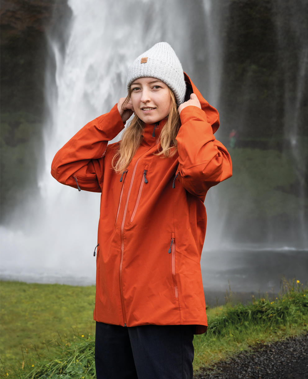 En person står foran et kraftfuldt vandfald med tæt tåge og vandstænk. Hun bærer en lysgrå strikhue og en kraftig orange regnjakke; hætten løftes let, mens hun justerer den med begge hænder. Baggrunden viser frodigt grønt landskab og en våd grussti. Stemningen er frisk og eventyrlig, med fokus på naturens rå energi og et roligt, forventningsfuldt ansigtsudtryk.