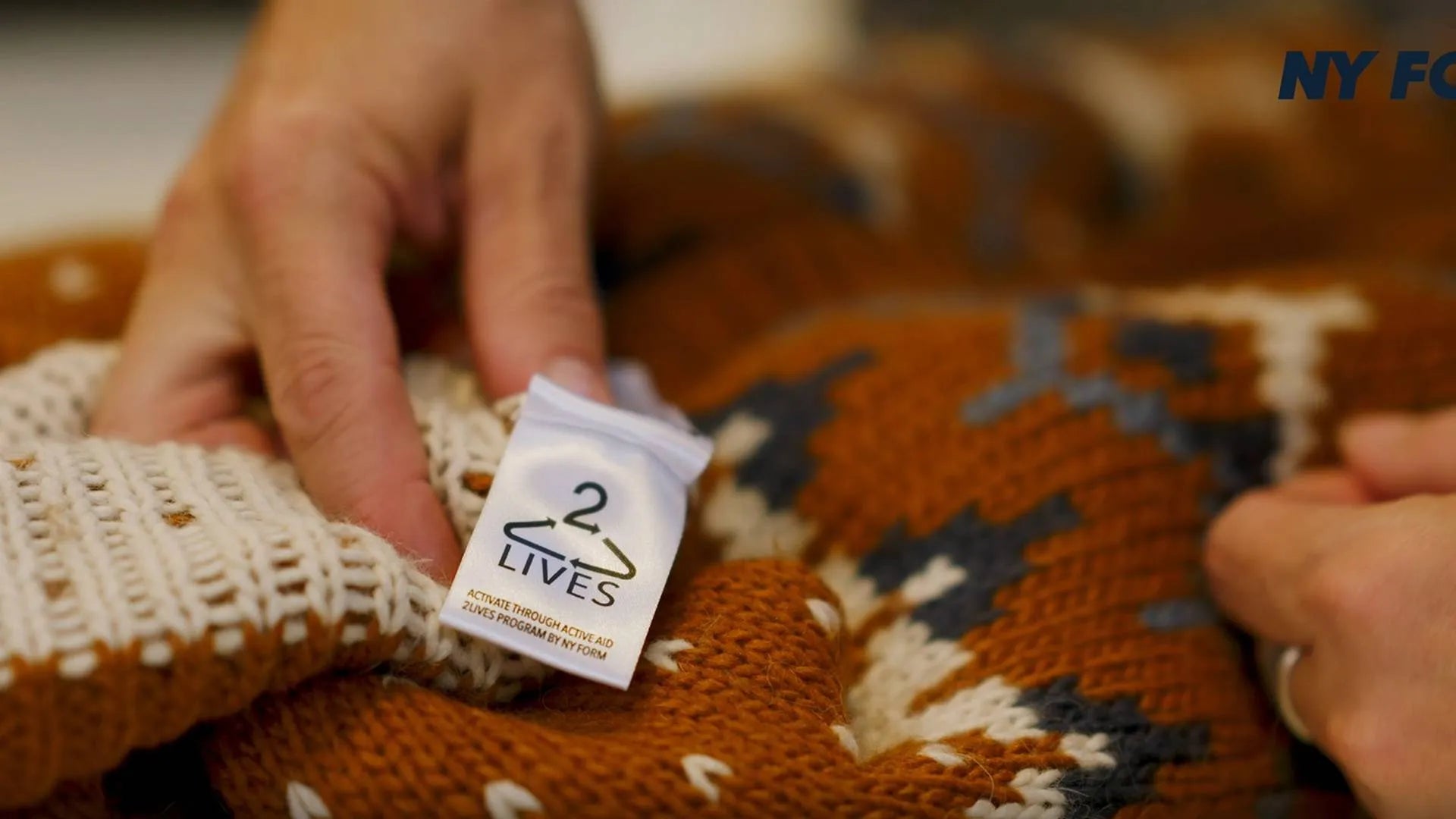 Billedet viser et nærbillede af hænder, der holder en varm, brun-orange strikket sweater med mønster. En hvid mærkeseddel med logoet '2 LIVES' trækkes frem, mens tekst og fold på stoffet vises tydeligt. Den bløde tekstur, de håndsyede detaljer og de kontrasterende farver skaber et hjemligt, håndlavet indtryk af tøj og omsorg. Det varme lys forstærker stemningen.