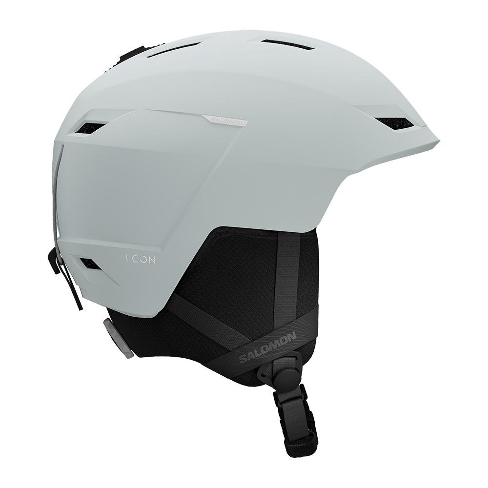 Et moderne, mat lysegråt skihjelm med strømlinet design og sorte, polstrede øreklaffer. Hjelmen har flere ventilationsåbninger, integreret skygge over panden og en justerbar hakestroppesystem med klikspænde. Brandnavnet er diskret trykt på siden, og formen signalerer både komfort og sikkerhed til alpine aktiviteter. Den minimalistiske æstetik gør den velegnet til både fritidsløbere og erfarne skiløbere.