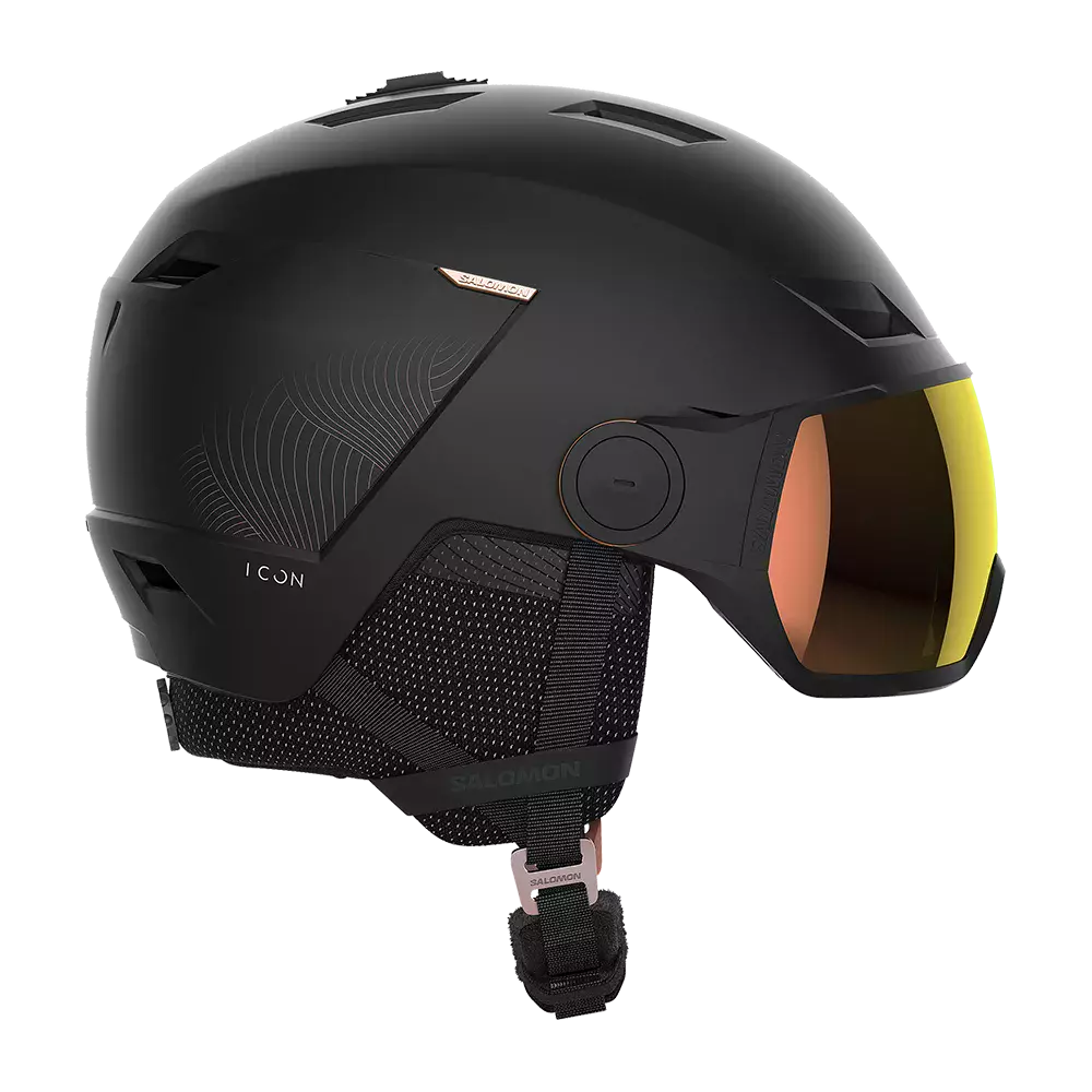 Mat sort skihjelm med indbygget visir og tonede linser giver et moderne og strømlinet udtryk. Hjelmen har flere ventilationsåbninger, polstrede ørepuder og en justerbar hakestroppes spænde for komfort og sikker pasform. Overfladen er mat med diskrete grafikdetaljer og et lille logo. Designet peger mod alpine vintersportsaktiviteter, hvor både beskyttelse, funktionalitet og stil prioriteres. Visiret beskytter mod sol og blænding samtidig med god udsyn.