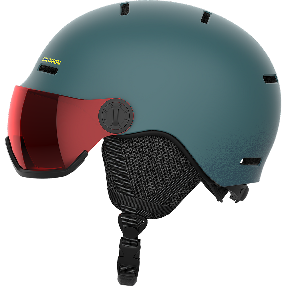 En moderne Salomon-skihjelm i mat blågrøn med integreret rødtonet visir, der dækker øjne og næse. Hjelmen har flere ventilationsåbninger, polstrede øreklapper i netmateriale og en justerbar hakestrop med sikkerhedsspænde. Den afrundede form og glatte overflade giver et stilrent udtryk, mens visiret beskytter mod blænding og vind, hvilket gør hjelmen velegnet til skiløb og snowboarding.