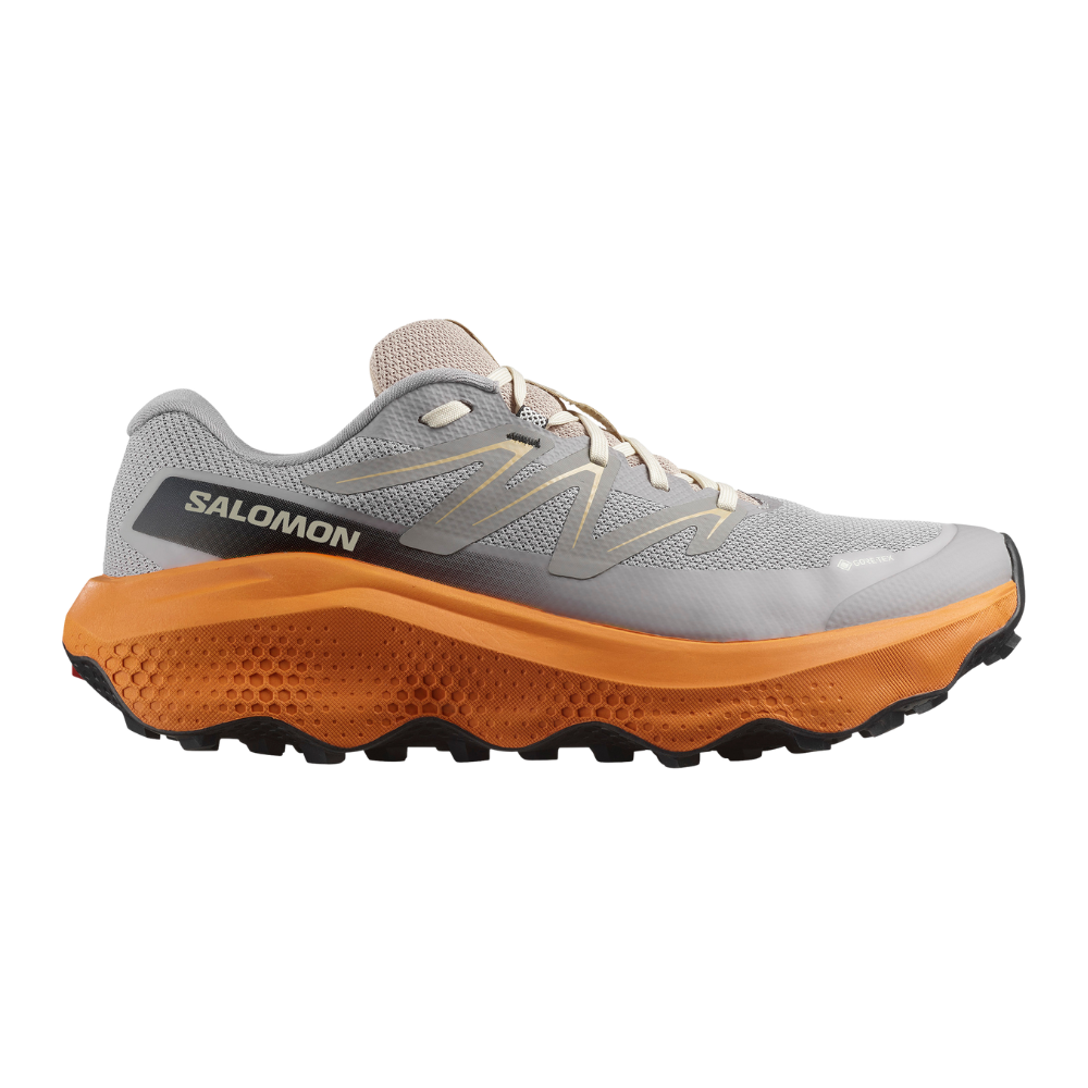 Et Salomon trail løbesko i profil; overdelen er lys grå mesh med diskrete beige detaljer og traditionelle snørebånd. Den markante orange mellemsål har kraftig dæmpning og honeycomb-tekstur, mens den sorte ydersål har aggressive mønstre for greb. Logoet "Salomon" ses på hælstykket, og en lille GORE-TEX-markering antyder vandtæt, åndbar membran. Skoen fremstår robust designet til ujævne stier og lange ture.