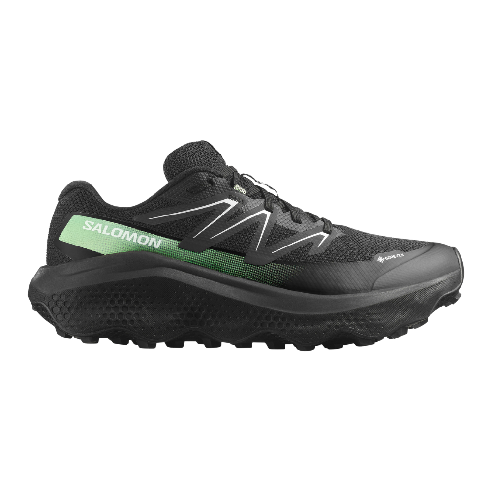 Et robust løbesko fra Salomon i sort med en grøn accent og tydeligt logo på siden. Skoen har et åndbart mesh-overmateriale, forstærkede syninger og Gore-Tex-mærkning, hvilket antyder vandtæthed. Den kraftige, aggressive ydersål og polstrede mellemsål giver godt greb og stødabsorbering til trail-løb. Et stilrent og funktionelt design, velegnet til ture i vådt og ujævnt terræn.