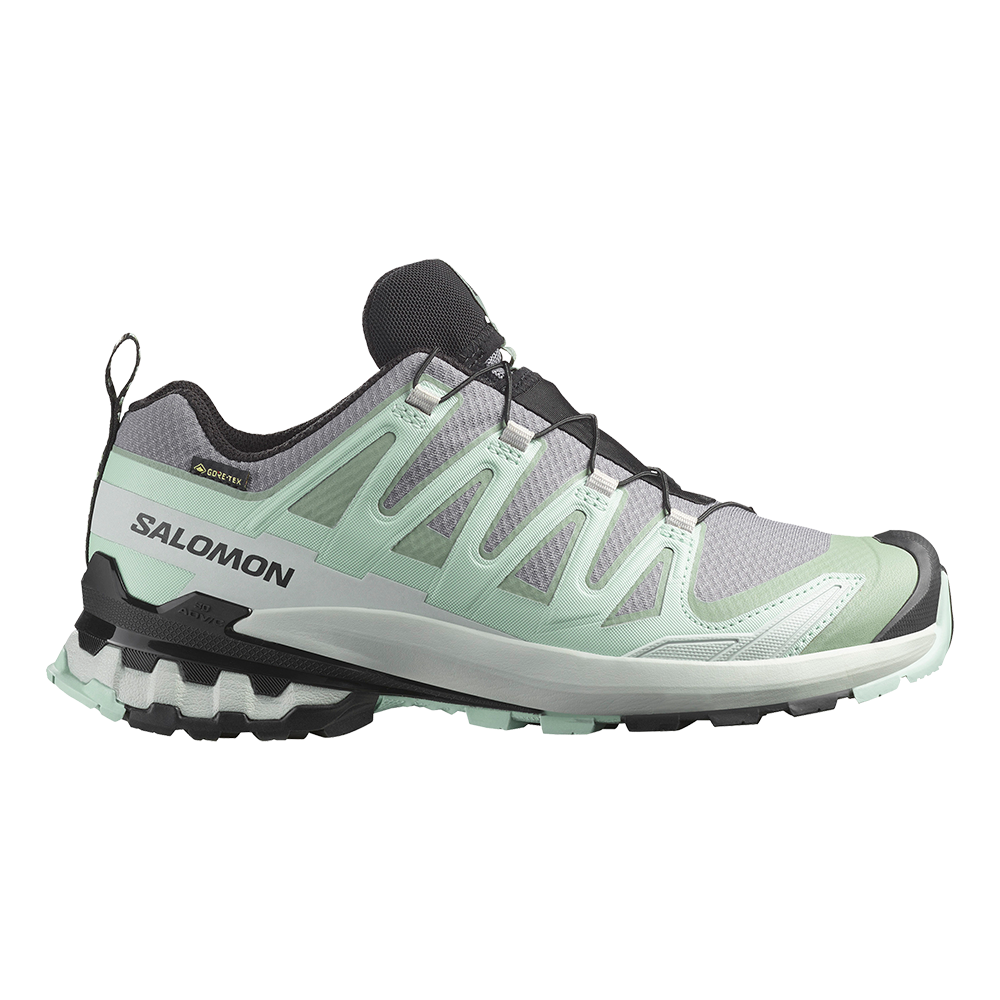 En lavt skåret Salomon trail-sko i mintgrøn, lys grå og sorte detaljer. Overdelen har profilerede syntetiske overlays og mesh, der giver støtte og åndbarhed, mens Gore‑Tex-mærket antyder vandtæt beskyttelse. Skoen har et hurtigt snøresystem, polstret hælkappe og en aggressiv, ruglet ydersål med kraftig slidbane for godt greb på mudrede stier. Designet virker let og teknisk til udendørs løb og vandring.