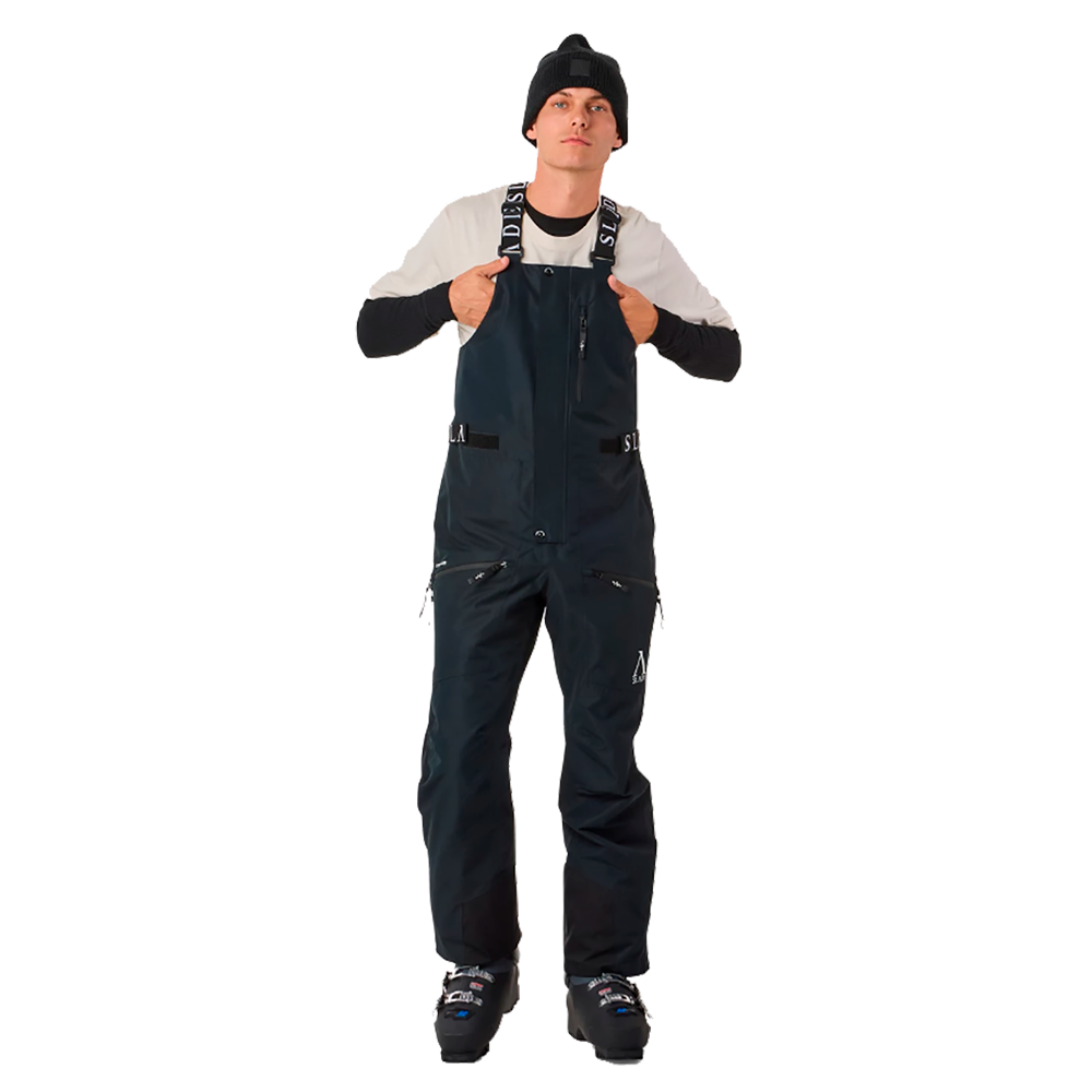 På billedet ses en ung mand iført Slade Aspen Bib Pants, som er designet til skisport. Disse mørkeblå skibukser fremstår med justerbare seler, der tydeligvis sikrer en behagelig pasform. Bukserne er udstyret med flere lynlåslommer, praktisk til opbevaring under aktivitet. Samlet set kombinerer bukserne funktionalitet med et moderne snit, perfekt til den stilbevidste skiløber.
