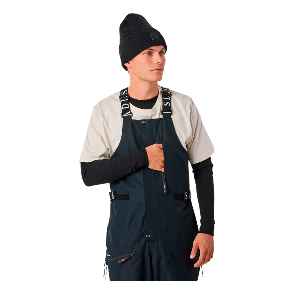 En ung mand demonstrerer Slade Aspen Bib Pants for herrer, som er ideelle skibukser til vintersport. Bukserne er dyb marineblå med justerbare skulderstropper, der er mærket med Slade-logoet. De har flere funktionelle lommer og er designet til at tilbyde både komfort og holdbarhed under skiløb eller snowboarding. Han bærer en sort strikhue, hvilket supplerer det stilfulde, sporty udtryk.