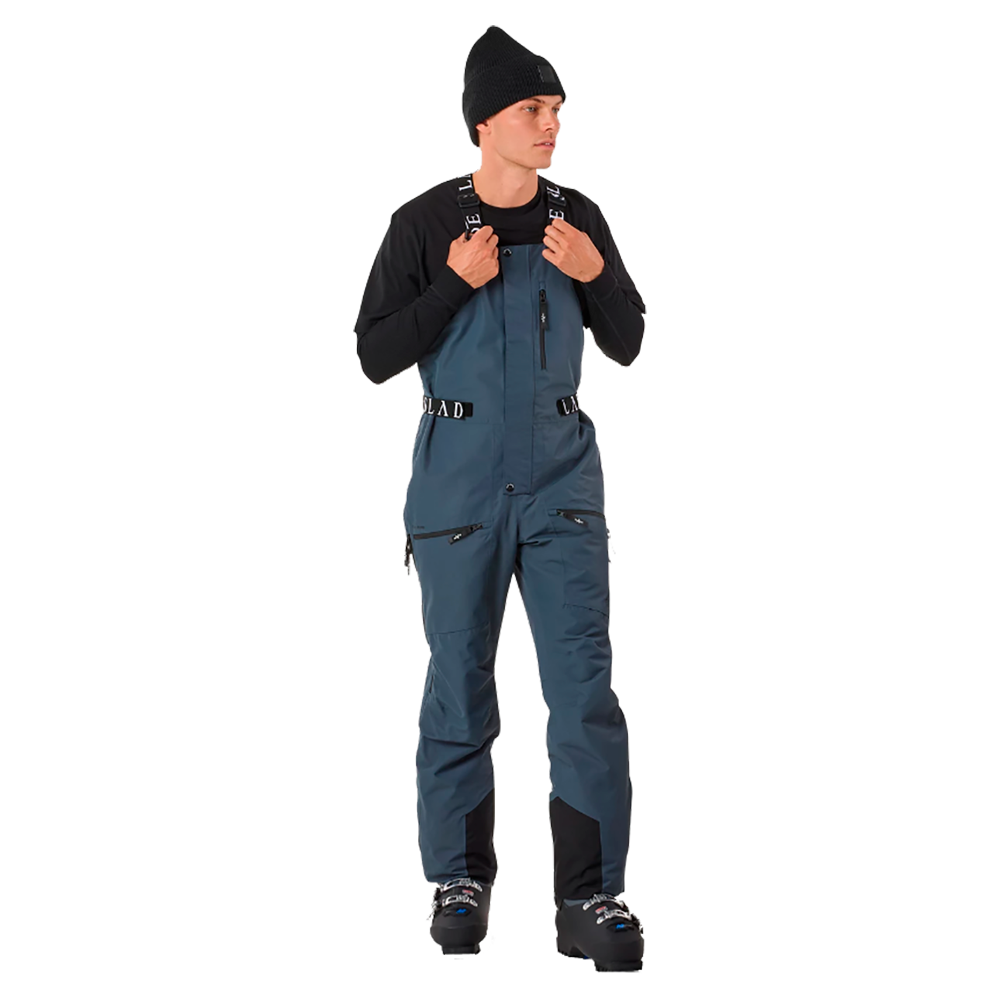 På billedet ses en ung mand, der præsenterer Slade Aspen Bib Pants, som er designet til mænd. Buksen har justerbare seler, flere funktionelle lommer, og den blå farve fremhæver et moderne snit. Skibukserne fra Slade kombinerer stil med praktiske detaljer, hvilket gør dem ideelle til skisport og vinteraktiviteter. Detaljerne og kvaliteten sikrer holdbarhed og komfort i koldt vejr.