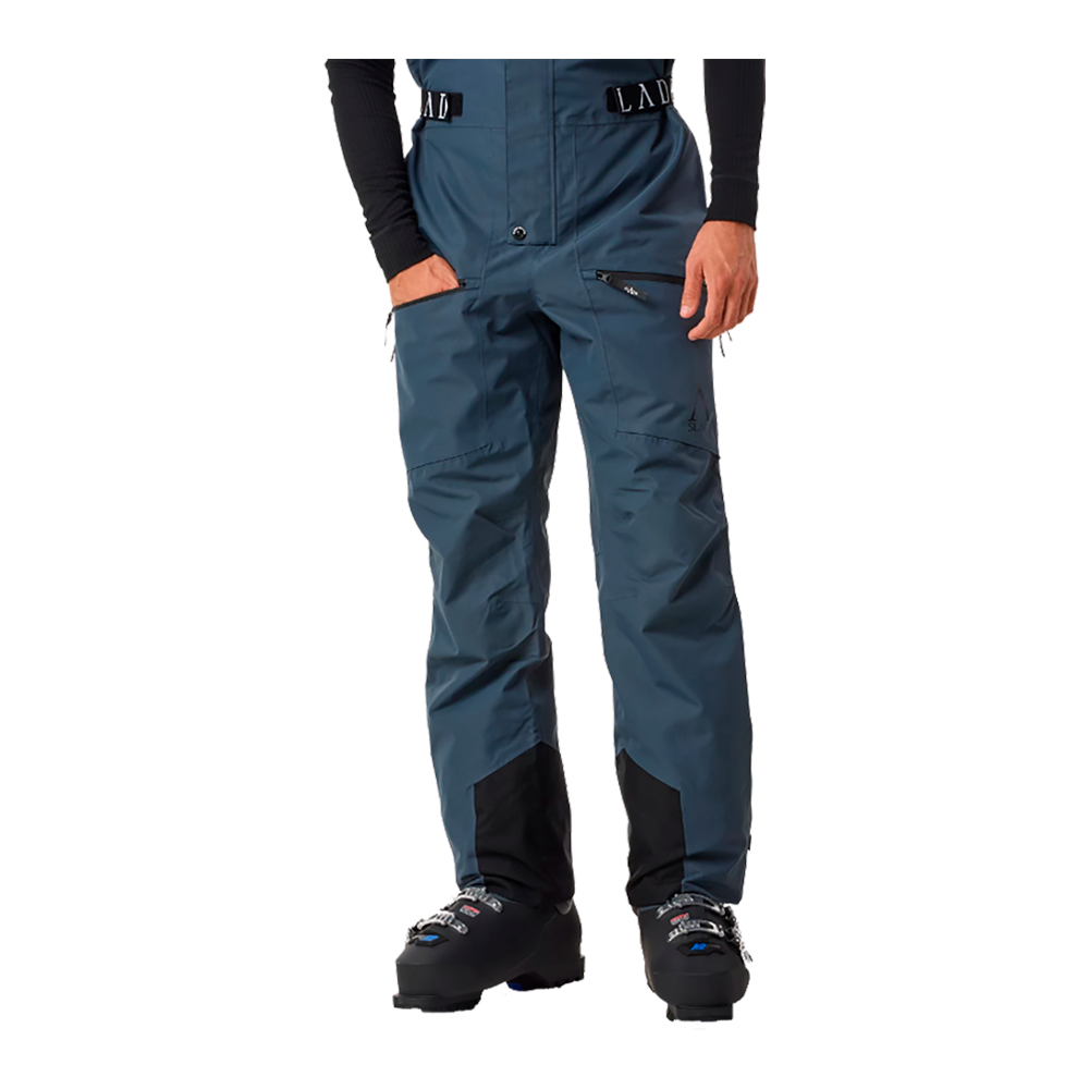 Billedet viser en ung mand, som står med et selvsikkert udtryk og iført Slade Aspen Bib Pants. Disse herre skibukser fra Slade er designet til at give optimal komfort og holdbarhed under vinteraktiviteter. Bukserne er blå med justerbare seler og flere lynlåslommer, ideelle til sikker opbevaring. En sort hue og skiferiefodtøj fuldender hans udstyr, hvilket fremhæver buksernes fleksibilitet og stil.