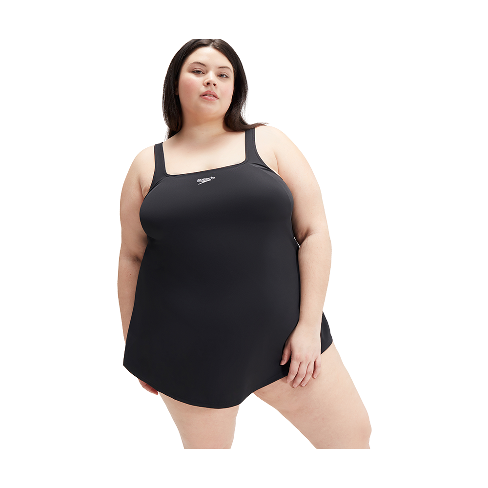 På billedet ses en person iført en enkel, sort badedragt med skørt (swimdress) og brede stropper. Modellen står i en selvsikker, afslappet positur mod en neutral, hvid baggrund. Badedragten har firkantet halsudskæring og et lille logo foran, hvilket giver et klassisk og funktionelt udtryk. Billedet udstråler komfort, inkludering og en tidløs stil, velegnet til både svømning og afslapning ved vandet.