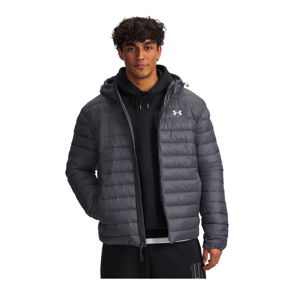 En ung person står afslappet i en mørkegrå, polstret jakke med hætte og tydeligt logo på brystet. Under jakken ses en sort hættetrøje med snøre og et hvidt lag nederst, mens hænderne hviler i lommerne. Jakken har vandrette syninger og en afslappet pasform, som giver et sporty og funktionelt udtryk, velegnet til hverdag og køligt vejr.