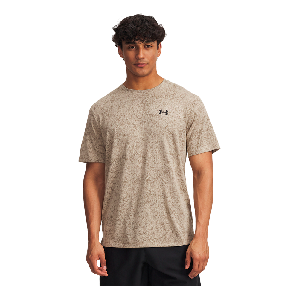 En ung mand står afslappet og ser direkte mod kameraet iført en beige, let meleret T-shirt med et diskret Under Armour-logo på brystet. Kort, krøllet mørkt hår og neutral mimik giver et naturligt indtryk. T-shirten har løs pasform og korte ærmer, kombineret med mørke shorts. Billedet fremhæver et simpelt, sporty og hverdagspræget look med fokus på komfort.