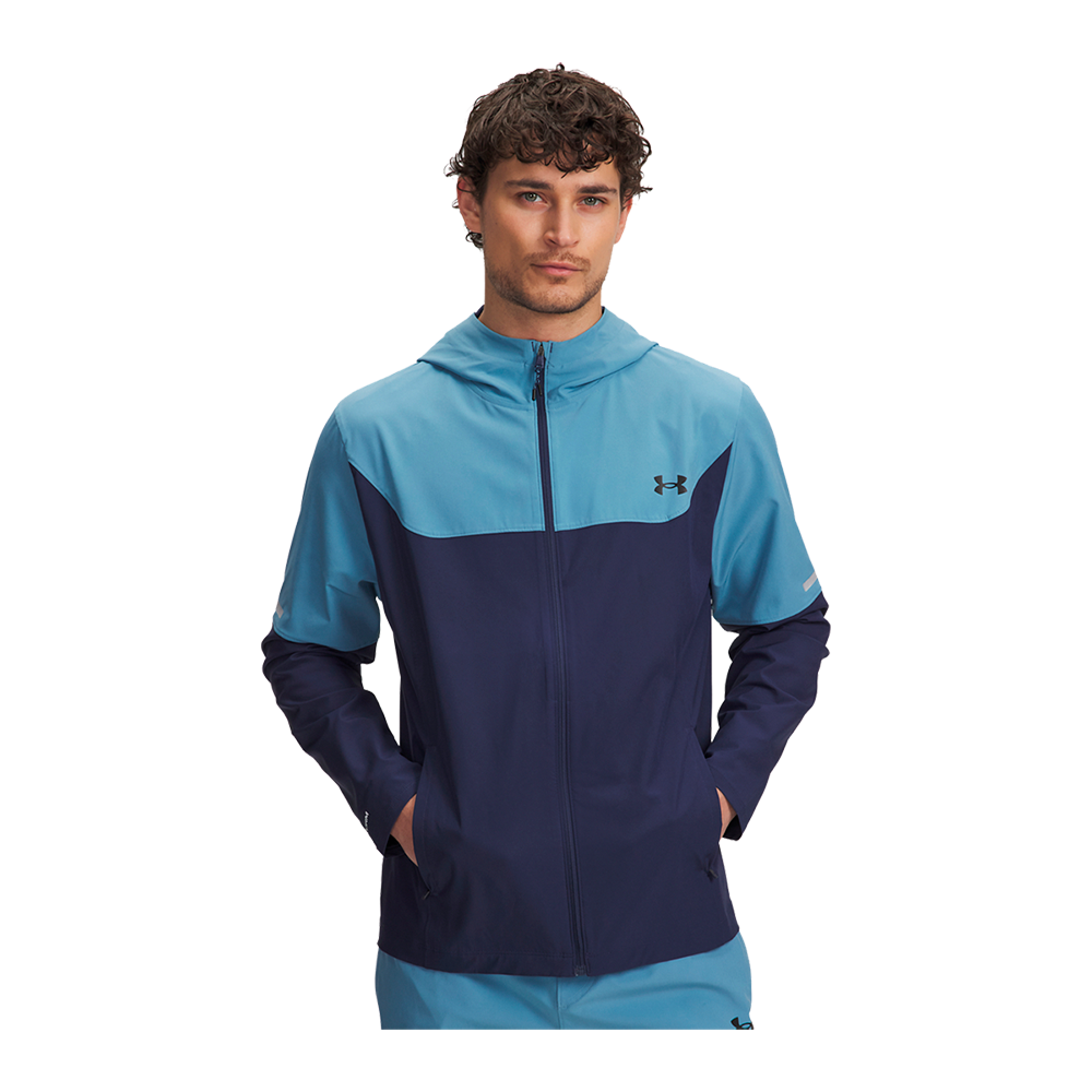 En mand iført en tofarvet træningsjakke med hætte står med hænderne i lommerne. Jakken er lyseblå øverst og marineblå nederst med en synlig lynlås og et lille logo på brystet. Han har kort krøllet hår og et neutralt, fokuseret udtryk. Stilen fremstår sporty og afslappet, velegnet til løb eller fritid, med rene linjer og funktionelle detaljer.