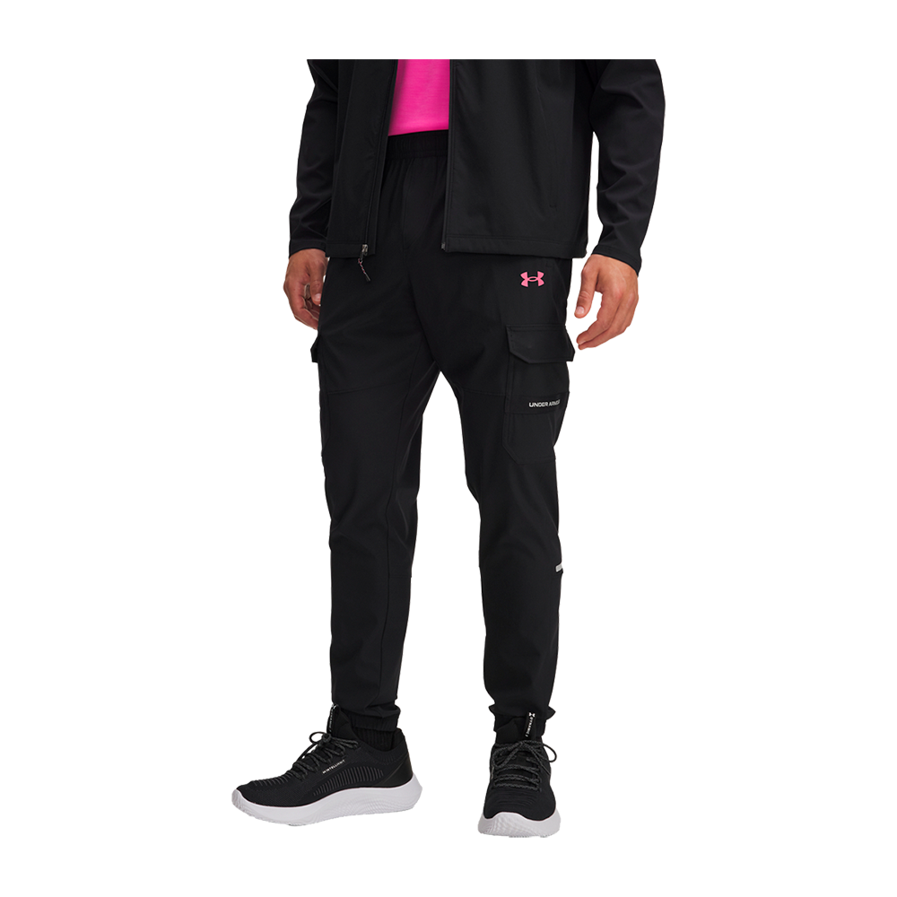 Billedet viser personens underkrop iført sorte træningsbukser med cargo-lommer og slank, tapered pasform. Bukserne har elastisk talje, lynlåsdetaljer og et lyserødt Under Armour-logo på låret, kombineret med sorte sneakers med hvide såler. En sort jakke og en pink top skimtes ovenover. Sættet fremstår moderne og funktionelt — velegnet til træning eller afslappet hverdagsbrug. Detaljer som lommelåg og små refleksstriber øger både stil og funktionalitet.