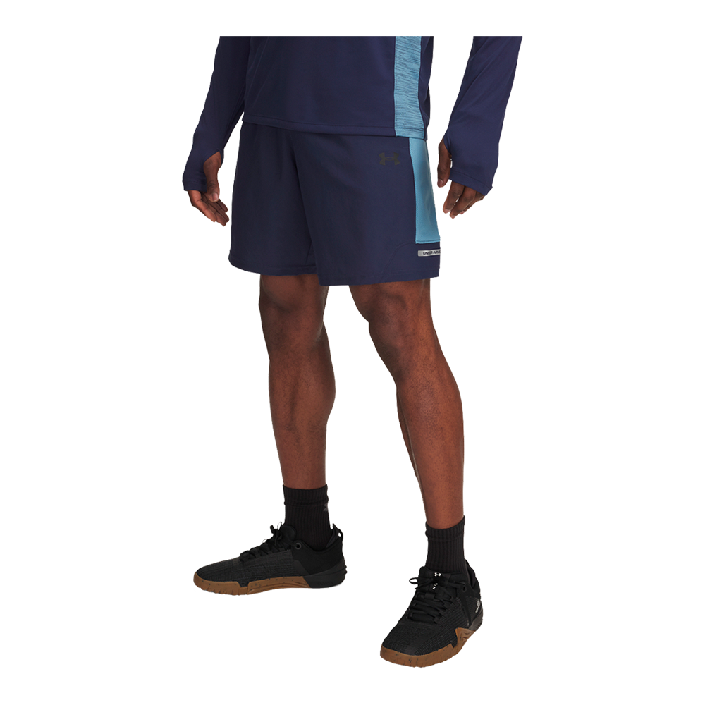 Dette billede viser underkroppen af en person iført sportstøj: navyblå træningsshorts med en lyseblå sideløsning og matchende langærmet overdel med tommelhul. Shortsene har et diskret logo og en løs pasform, der giver bevægelsesfrihed. Personen bærer sorte træningssokker og sorte sneakers med lysebrun gummisål. Samlet signalerer outfittet en funktionel og stilren træningsstil til indendørs eller udendørs aktivitet.