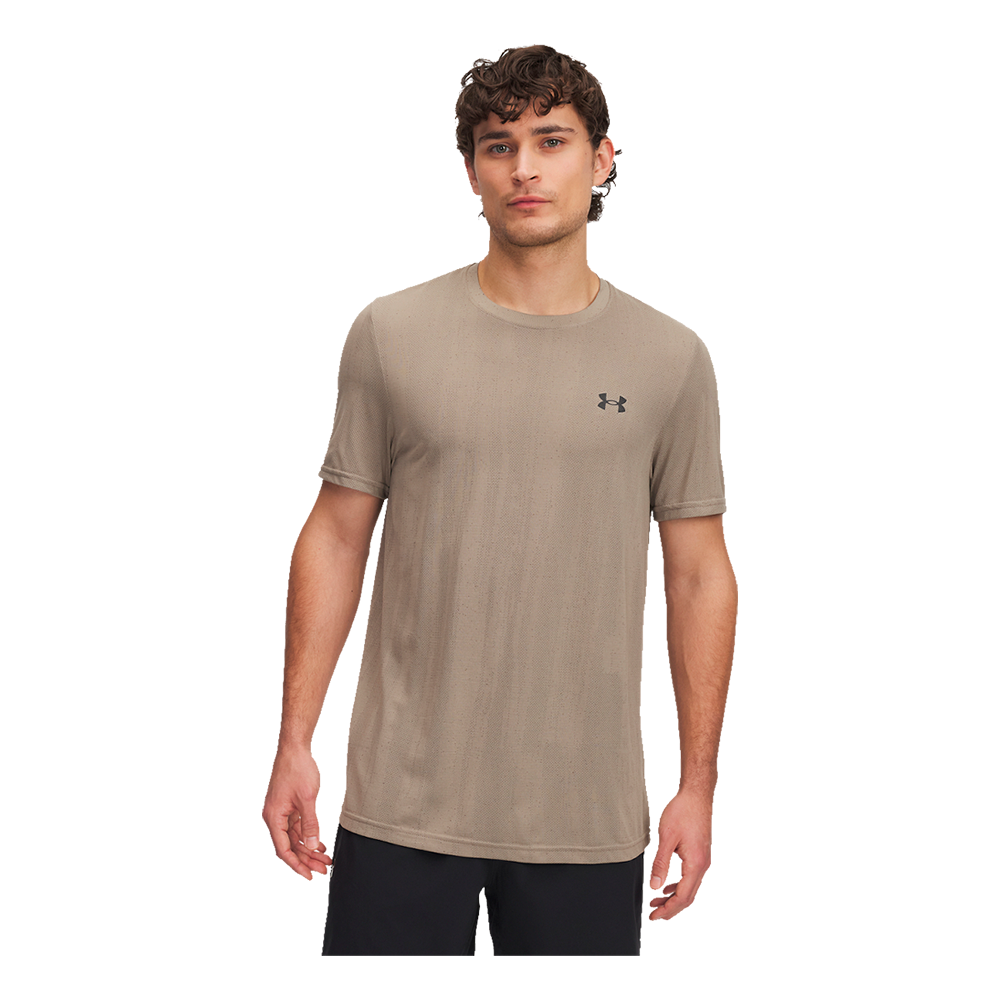 En ung mand står i en neutral studiobelysning iført en lys beige Under Armour t‑shirt med kortærmer og rund hals. T‑shirten har et diskret logo på brystet og et let, åndbart materiale med små perforeringer, ideel til træning. Modellen har afslappet kropsholdning, mørkt krøllet hår og sorte træningsshorts. Billedet er enkelt og fokuserer på tøjets pasform og funktionalitet.