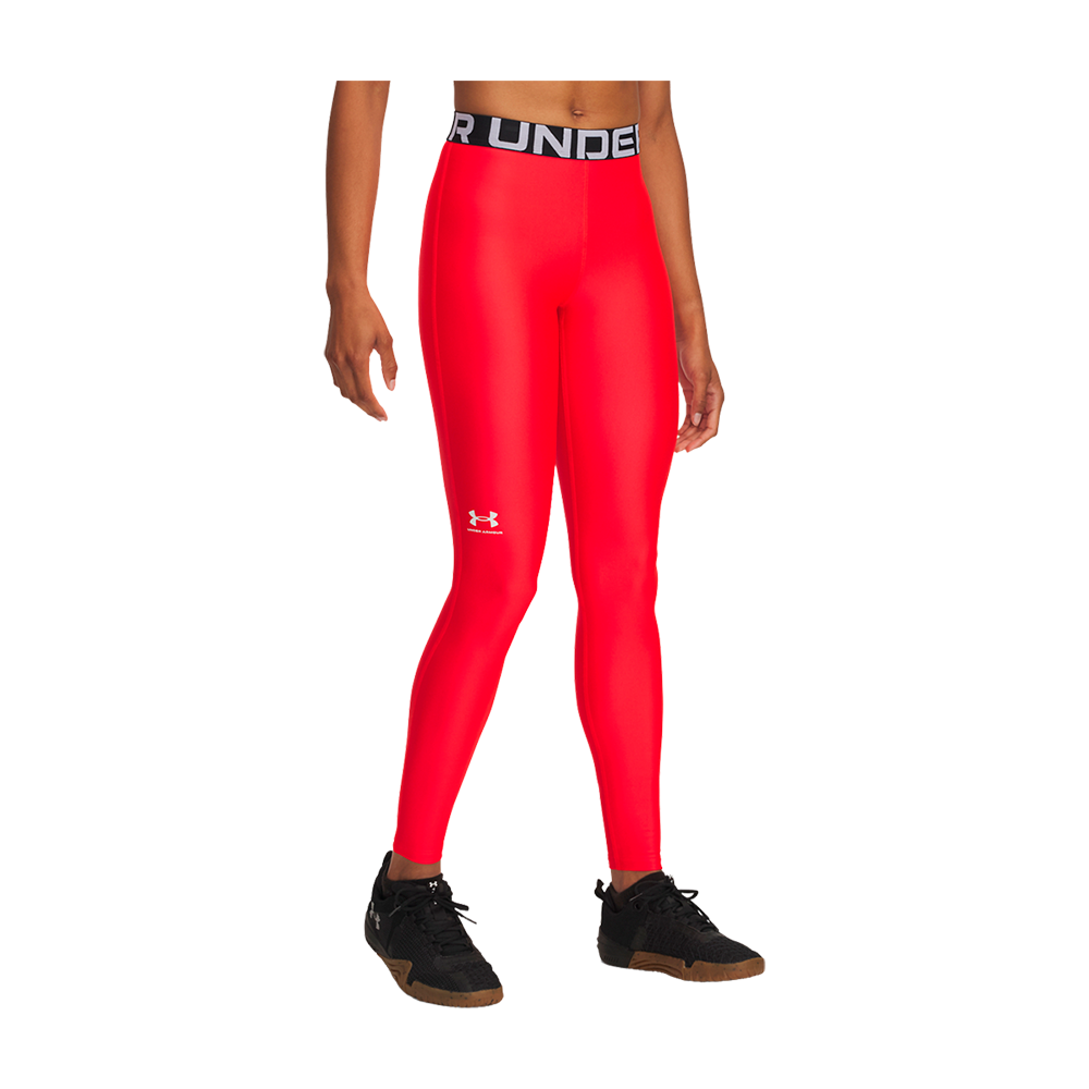 En model bærer et par skinnende, højttaljede træningsleggings i intens rød farve med en bred, sort elastik i taljen markeret af "UNDER ARMOUR"-tekst. Leggingsene har tæt, kropsnær pasform og et lille hvidt logo på låret. Kombinationen med sorte træningssneakers giver et sporty, funktionelt udtryk, velegnet til fitness, løb eller hverdagsaktiviteter, hvor bevægelsesfrihed og synlig stil prioriteres.