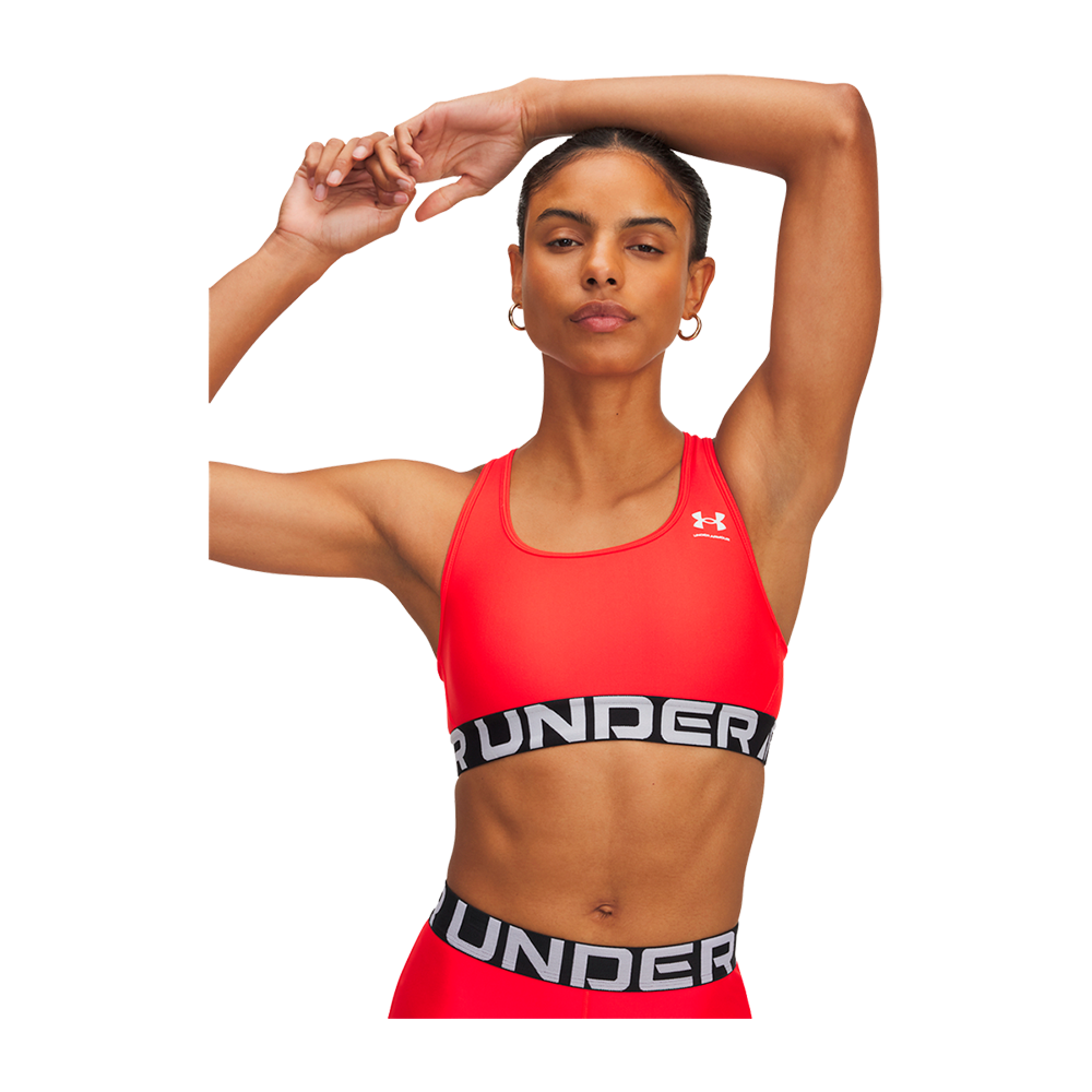 En atletisk kvinde iført en klar rød sports-bh og matchende træningsbunde står i en selvsikker positur med armene løftet over hovedet. Hendes muskuløse overarme og veltrænede mave er tydelige, og hun bærer en elastisk linning med synligt logo. Hun har samlet hår, diskrete øreringe og et fokuseret udtryk, der signalerer styrke, koncentration og sportslig selvsikkerhed.