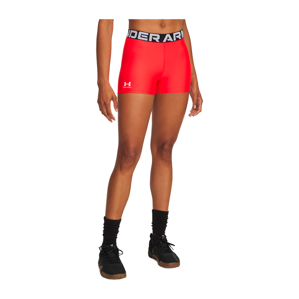 En sportslig scene viser en person iført tætsiddende, røde træningsshorts med en bred sort linning prydet af 'UNDER ARMOUR'-logoet og et lille hvidt mærke på låret. Modellen står afslappet med let adskilte ben, veltrænede ben og brun hudtone. Outfit'et kompletteres af sorte mellemhøje sokker og sorte sneakers, hvilket giver et enkelt og atletisk look velegnet til træning eller restitution.