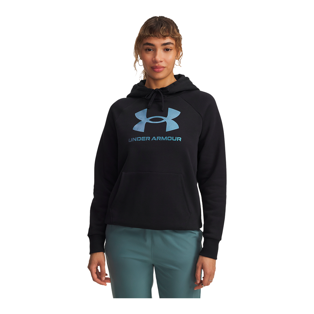 En person står afslappet i en sort hættetrøje fra Under Armour med et stort lyseblåt logo på brystet. Trøjen har snøre i hætten, kængurulomme og ribkant ved ærmerne. Personen bærer grønne-turkise bukser og har naturligt, bølget hår opsat med pandehår. Udtrykket er roligt og neutralt; billedet er rent og fokuserer på sportstøjets pasform, detaljerne og materialets struktur.