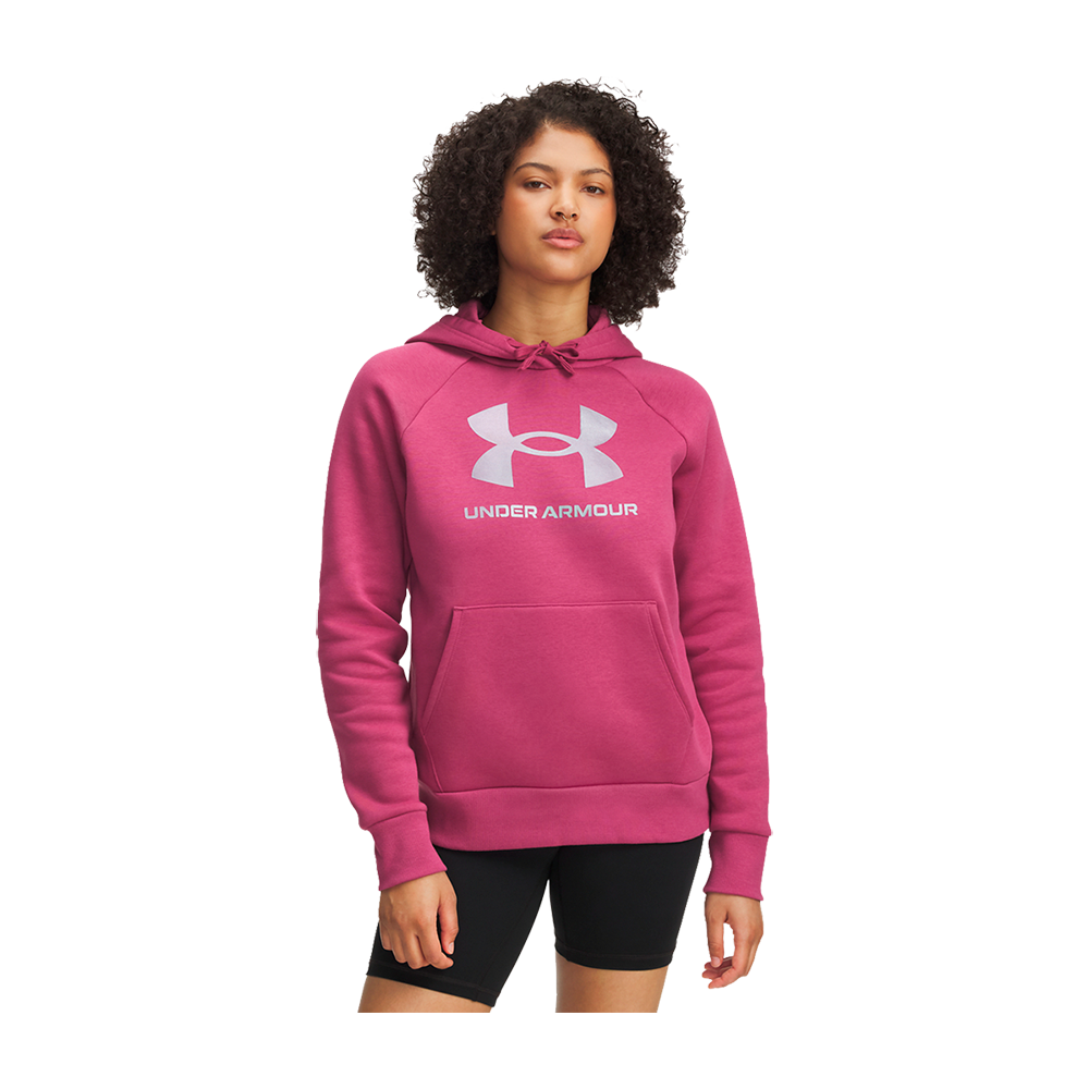 En ung kvinde står afslappet i en knaprosa hættetrøje med stort hvidt Under Armour-logo og kængurulomme. Trøjen har snører i hætten og lange raglanærmer. Hun bærer korte, sorte træningsshorts, har krøllet hår og et roligt, selvsikkert udtryk. Looket virker sporty og komfortabelt, egnet til både træning og afslappet hverdag.