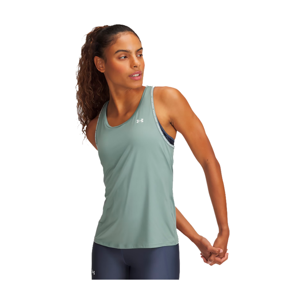 En kvinde står i en afslappet strækøvelse, med blikket vendt mod siden. Hun bærer en mintgrøn træningstop med Under Armour-logo og mørkeblå træningstights. Håret er trukket tilbage i en hestehale, og kroppen udstråler ro og fokus. Baggrunden er neutral, hvilket fremhæver tøjets klare farver og den atletiske holdning. Billedet virker både energisk og harmonisk, velegnet til sport og fritid.