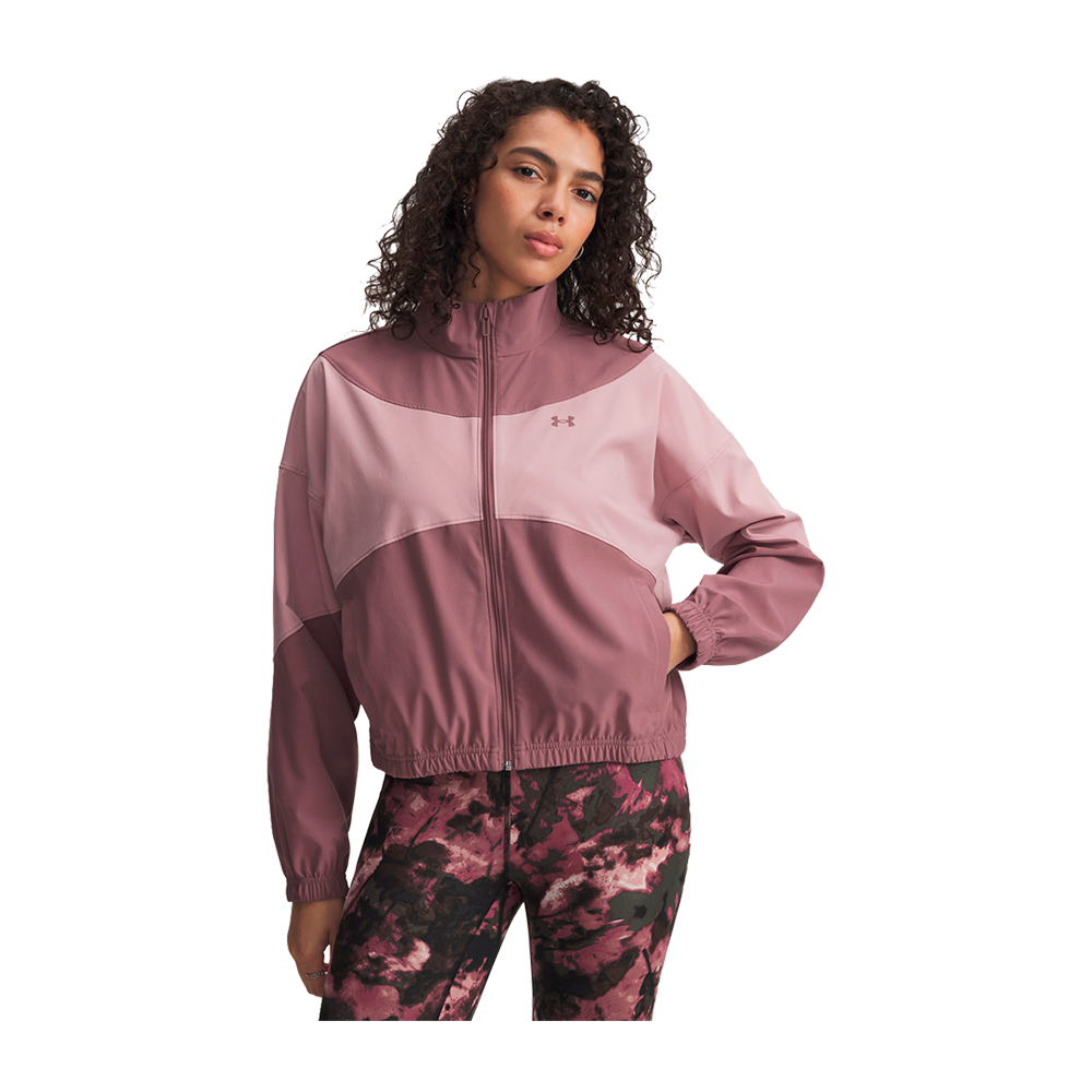 En person står i en afslappet positur iført en tofarvet lyserød og mørkrosa vindjakke med høj krave, fuld lynlås, elastisk kant og et diskret logo på brystet. Jakken er kombineret med matchende træningsleggings i mørke rosa og sort i et abstrakt marmoreret mønster. Personen har krøllet hår og en rolig, selvsikker mine, med den ene hånd i lommen.