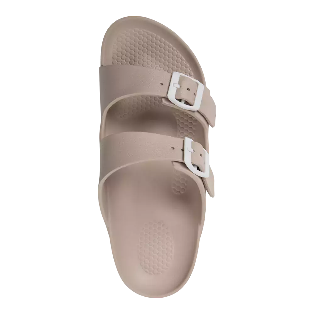 Billedet viser en enkel, beige sandal set oppefra. Den har to brede remme med hvide spænder og en blød, tekstureret sål med mønster for bedre greb. Sålen er formet ergonomisk med let hævning ved hælen og afrundet tåparti. Materialet ser ud til at være let og vandafvisende, hvilket gør sandalen praktisk til hverdag, strandture og afslappede sommerdage.
