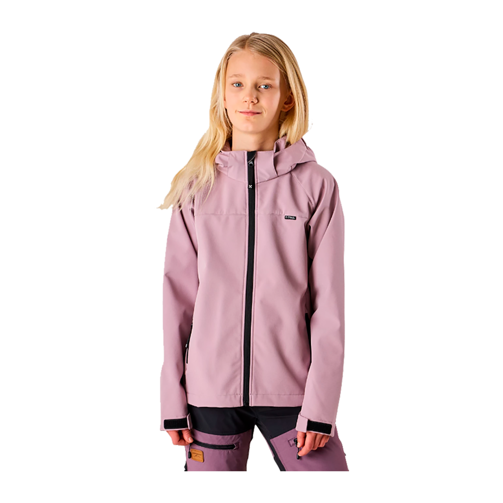 En ung pige står afslappet i en lys lilla softshelljakke med hætte og sort kontrastlynlås. Jakken har justerbare ærmer med velcro og sidelommer, og hun bærer matchende bukser med lynlåslommer. Hendes lange, lyse hår er løst omkring skuldrene, og ansigtsudtrykket er roligt og selvsikkert. Baggrunden er neutral, hvilket fremhæver det funktionelle, minimalistiske udendørslook.