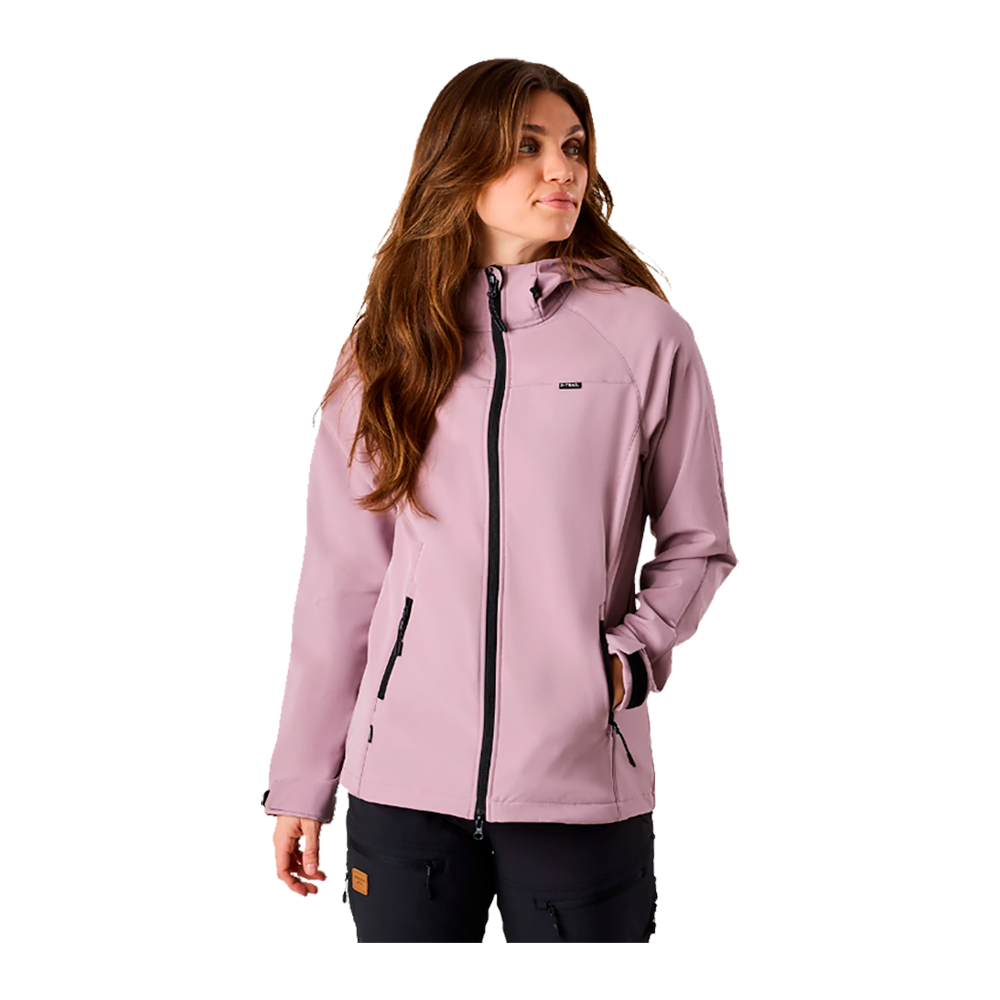 En kvinde iført en lyserosa softshell-jakke står i afslappet positur med hænderne i lommerne, vendt let mod højre. Jakken har sort lynlås og praktiske sidelommer, og sidder tæt ved hoften. Hendes langt, brunt hår falder løst over skuldrene, og hun giver udtryk for rolig opmærksomhed. Billedet fremhæver jakken som moderne, funktionel og velegnet til udendørs brug.