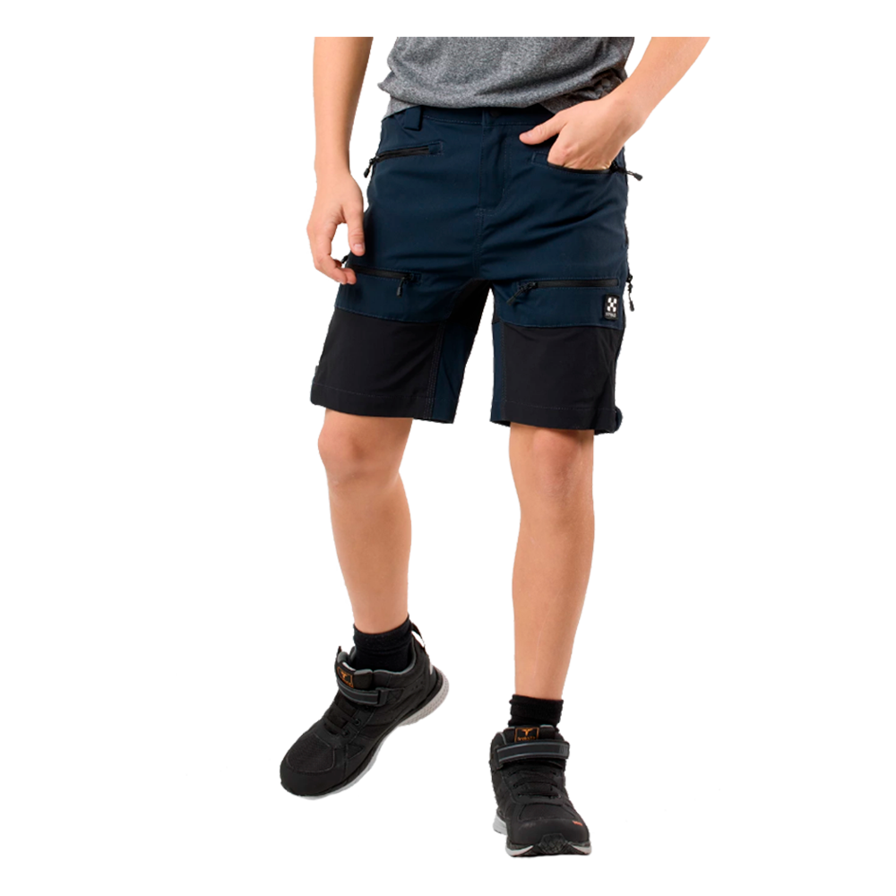 En person er afbildet fra livet og ned iført mørkeblå og sorte cargo-shorts med flere lynlåslommer, en grå T-shirt er delvist synlig. Shortsene har praktiske lommer omkring lårene og en kontrast i stoffet. Personen bærer sorte træningssko med velcro-stropper og sorte sokker. Billedet fremhæver et funktionelt, sporty og afslappet hverdags- eller arbejdstøjslook.