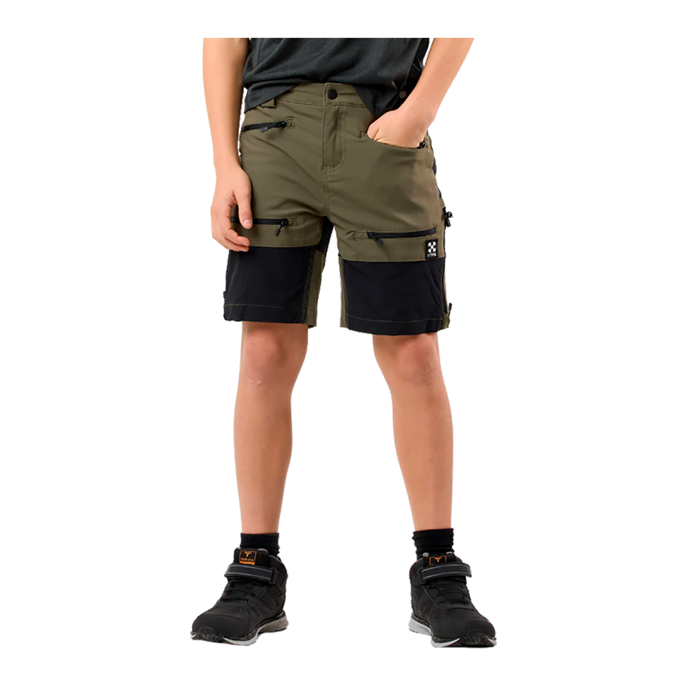 Billedet viser et par funktionelle, tofarvede shorts i olivengrøn og sort. Shortsen har flere lynlåslommer på lårene og ved hofterne, forstærkede sorte paneler forneden og en komfortabel pasform til knæet. Modellen står i afslappet stilling med hænderne i lommerne og bærer sorte ankelsokker og robuste, sorte sneakers med velcro, hvilket understreger et praktisk og sporty udtryk.