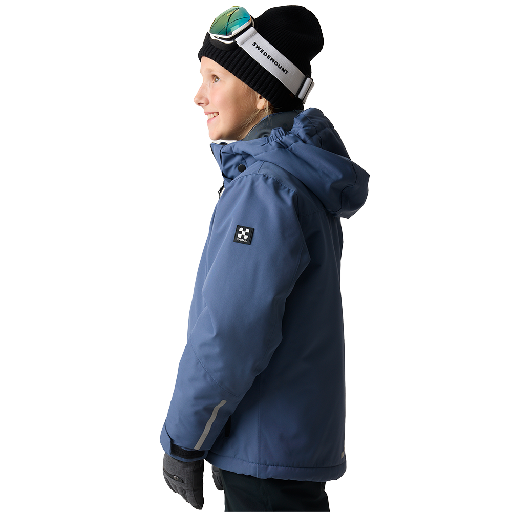 Billedet viser et barn i profil iført en tyk, blå vinterjakke med hætte, der ser glad og forventningsfuld ud. Jakken har en diskret logo-patch på ærmet og reflekterende detaljer på underarmen. Barnet bærer en sort hue og skibriller anbragt oven på hovedet samt varme handsker, hvilket giver indtryk af parathed til en dag i sneen eller udendørs aktiviteter i koldt vejr.