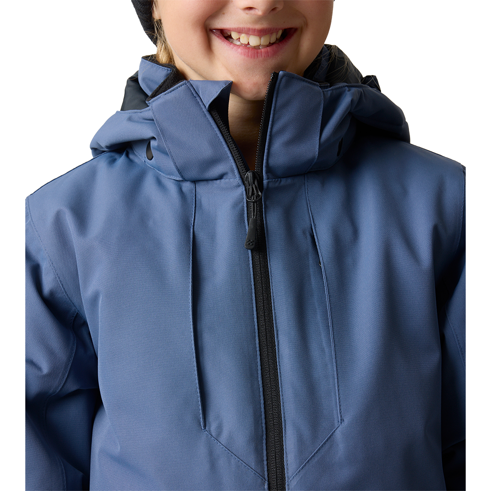 Et nærbillede viser en smilende person iført en matblå, vandafvisende jakke med høj krave og tilhørende hætte. Den sorte midterlynlås er trukket helt op til hagen, og stoffets sømme og paneler er tydelige. I toppen ses kanten af en sort hue, mens personens smil giver billedet et varmt og imødekommende præg. Jakken fremstår teknisk og funktionel til koldt vejr.