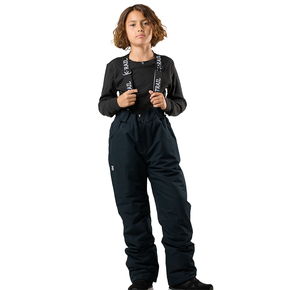 Et barn står oprejst iført mørke, vandtætte skibukser med seler og en enkel, langærmet sort trøje. Bukserne sidder løstsiddende med forstærkede knæ og elastisk talje, velegnet til udendørs leg i koldt vejr. Personen har kort til skulderlangt, krøllet hår og et beslutsomt, neutralt udtryk, og poseringen viser funktionelt tøj designet til bevægelse og beskyttelse.