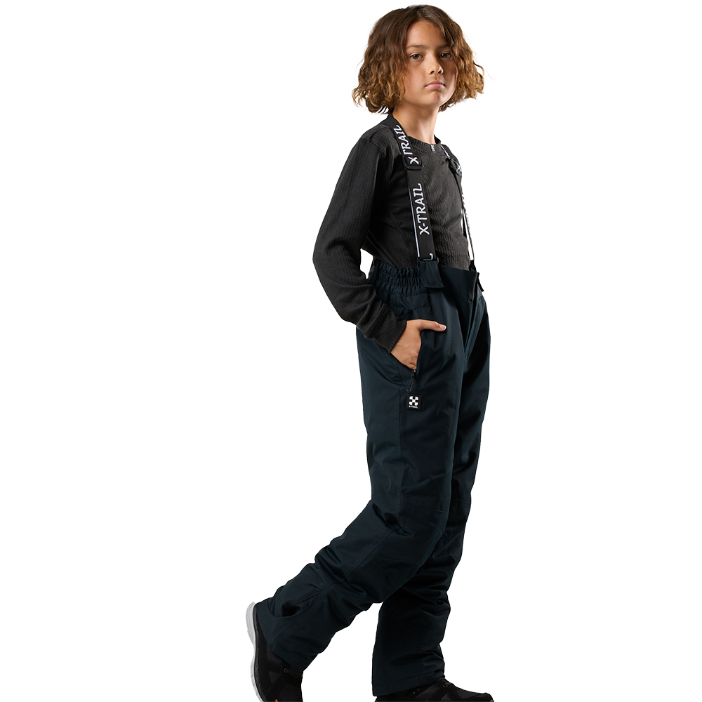 Et ungt model står i profil med hånden i lommen og et selvsikkert udtryk. Personen bærer mørke, vandtætte skibukser med elastisk talje og brede seler med teksten "X-TRAIL", kombineret med en sort, ribstrikket langærmet trøje og sorte sko. Tøjet giver et praktisk og sporty udtryk, velegnet til udendørs aktiviteter i koldt vejr.