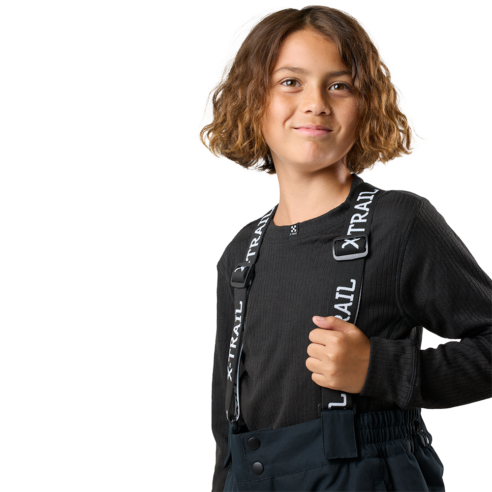 Et portræt af en ung person med skulderlangt, krøllet brunt hår og et mildt smil. Personen har en sort, ribstrikket bluse og sorte bukser med brede stropper, hvor teksten X-TRAIL ses. Billedet er taget mod en lys, neutral baggrund; kompositionen fokuserer på overkroppen og understreger et afslappet, selvsikkert udtryk samt praktisk, sporty påklædning.