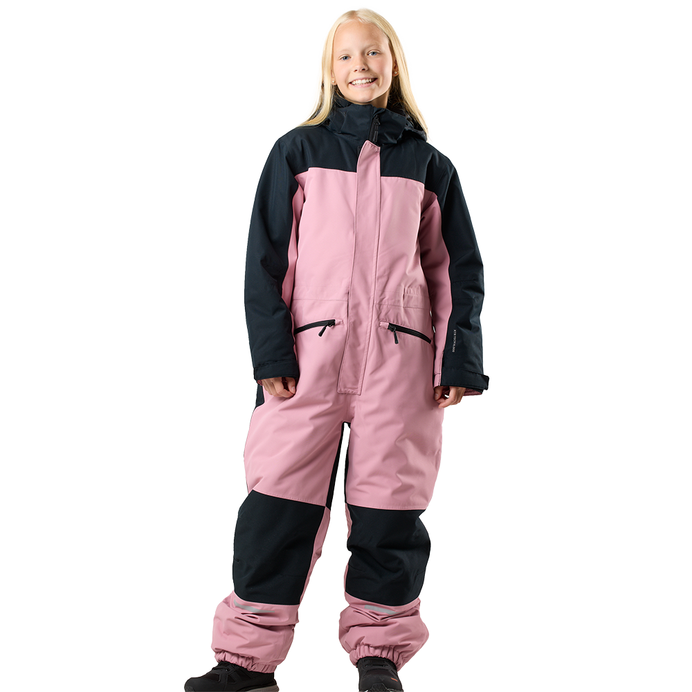 Billedet viser en smilende pige iført en lyserød og sort skidragt/overall. Dragten har høj krave og hætte, lange ærmer med velcrolukning, to forlommer med lynlås og forstærkede knæpartier i sort stof. Buksebenene er elastiske ved anklerne, og hun står i sorte støvler på en neutral, lys baggrund. Udtrykket er glad og afslappet, klar til udendørs aktiviteter i koldt vejr.