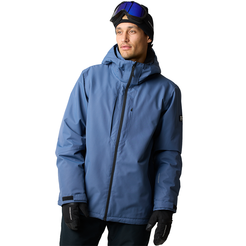 En ung mand iført en blå, vandtæt vinterjakke med høj krave og hætte står let vendt mod højre. Han bærer en sort hue og skibriller placeret oven på panden samt sorte handsker. Jakken har en central lynlås og rene, funktionelle detaljer, der signalerer praktisk funktionalitet til ski eller andre udendørsaktiviteter i koldt vejr.