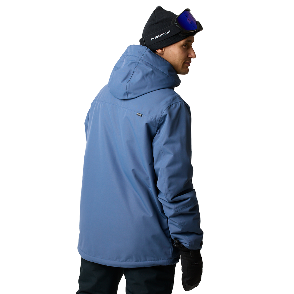 En mand iført en støvet blå skijakke vendt let væk fra kameraet. Jakken har hætte og et rent, funktionelt snit i vandafvisende materiale. Han bærer en sort hue med skibriller siddende ovenpå og sorte handsker, hvilket giver et praktisk vinterlook. Kombinationen signalerer robust, minimalistisk ski- eller friluftstøj designet til kolde forhold og bevægelsesfrihed på pisterne.