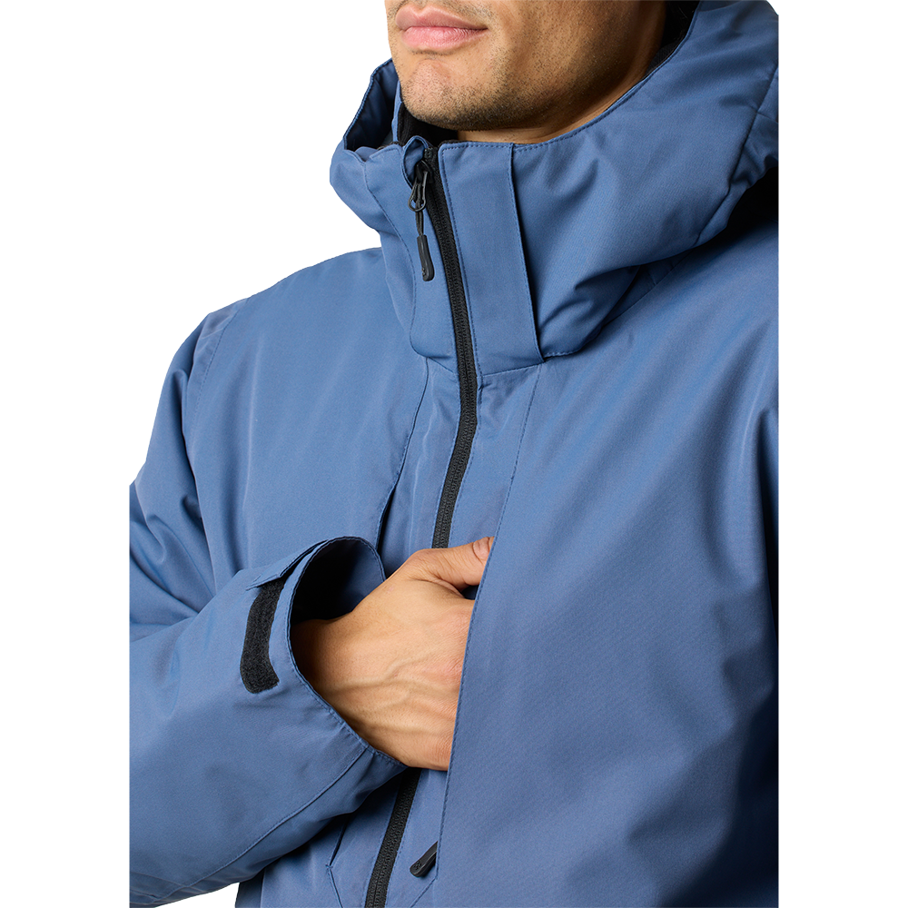 Nærbillede af en person iført en blå, polstret jakke med høj krave og hætte. Jakken har en central lynlås med stormflap, justerbar velcrolukning ved ærmet og en diskret bryståbning, hvor hånden glider ind. Stoffet ser mat og vandafvisende ud, hvilket giver et robust og funktionelt indtryk. Stilren og praktisk yderbeklædning, egnet til koldt og blæsende vejr.