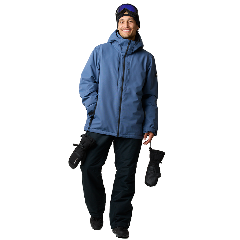 En ung person står afslappet iført en støvet blå vinterjakke med hætte, mørke skibukser og sorte skistøvler. Han smiler og har skibriller skubbet op på panden, mens han holder et par tykke sorte vanter i hånden. Tøjet ser teknisk og vandtæt ud, velegnet til skiløb eller friluftsliv, og hele udtrykket virker varmt, praktisk og klar til snefyldte eventyr.