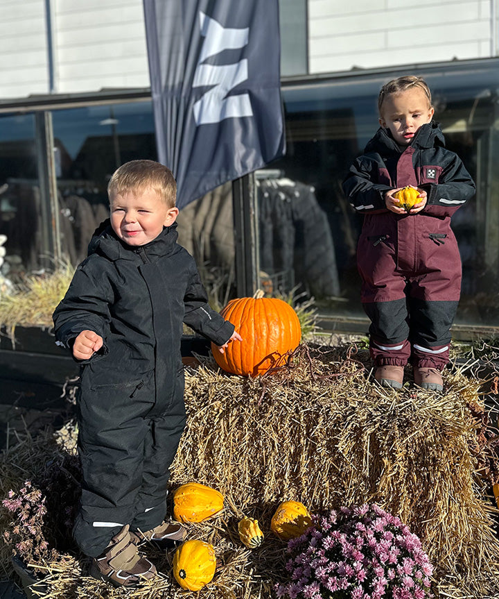 På billedet ses en ung pige iført X-TRAIL Cortina Overall Jr, en børneflyverdragt fra X-Trail. Dragten har en flot farvekombination af mørk bordeaux og marineblå, hvilket giver et stilrent og funktionelt udtryk. Modellen er designet til at modstå koldt vejr, med robuste materialer der holder barnet varmt og tørt. Derudover er flyverdragten forsynet med reflekser for sikkerhed i lav belysning, hvilket gør den ideel til vinterbrug i både skole og fritid.