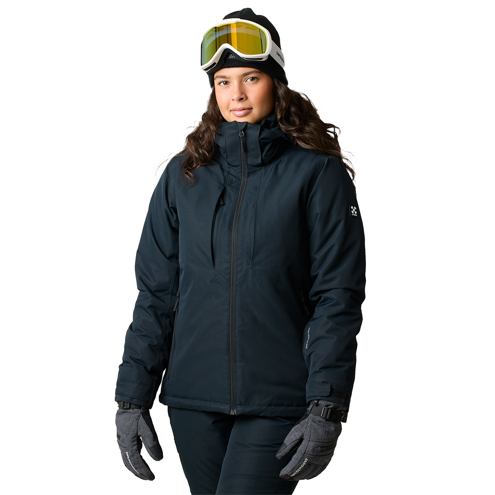 En ung kvinde står i en mørk vinterjakke med høj krave, varme handsker og en sort hue. Skibriller med gultonet linse hviler på huen, hvilket antyder ski- eller snowboardudstyr. Hendes holdning er afslappet og selvsikker, og tøjens funktionelle detaljer signalerer beskyttelse mod kulde og vind. Det enkle, lyse baggrundsudtryk fremhæver det praktiske, sporty outfit.