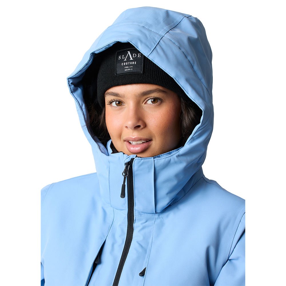 Billedet viser en person i en lyseblå, vatteret jakke med hætte trukket op. Under hætten ses en sort hue med synligt mærkefelt 'SLADE COUTURE'. Ansigtet er let smilende, med varm hudtone og naturligt makeup. Jakken har høj krave med sort lynlås og detaljer ved brystet, hvilket giver et moderne, funktionelt vinterlook, der signalerer både varme og stil.