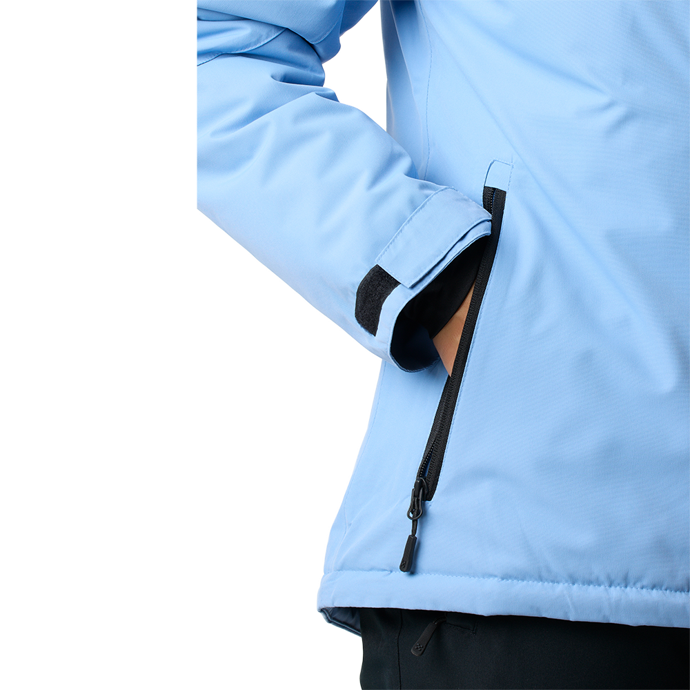 Nærbillede af en lyseblå jakke med fokus på ærmet og sidelommen. Jakken virker let polstret og har en vandafvisende lynlås, en velcrolukning ved håndleddet og sort indvendig kant, der skaber kontrast. En hånd ligger i lommen, hvilket viser praktisk pasform og funktionalitet. Designet fremstår enkelt og teknisk, velegnet til udendørs aktiviteter eller hverdag.