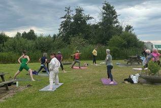 En gruppe mennesker deltager i en udendørs træningstime på en græsplæne. De står på yogamåtter med varierende farver og udfører udstrækningsøvelser under en grå, let overskyet himmel. Området er omgivet af buske og træer, og der står picnicborde og personlige ting i nærheden. Stemningen virker rolig og fokuseret, med god plads mellem deltagerne.