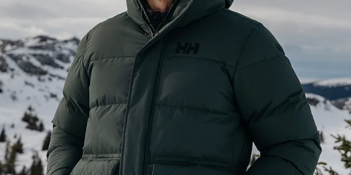 Mand i mørkegrøn Helly Hansen-vinterjakke og orange strikhue i et snedækket bjerglandskab.