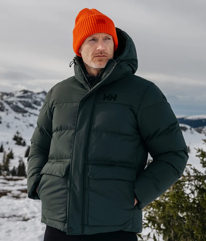 Mand i mørkegrøn Helly Hansen-vinterjakke og orange strikhue i et snedækket bjerglandskab.